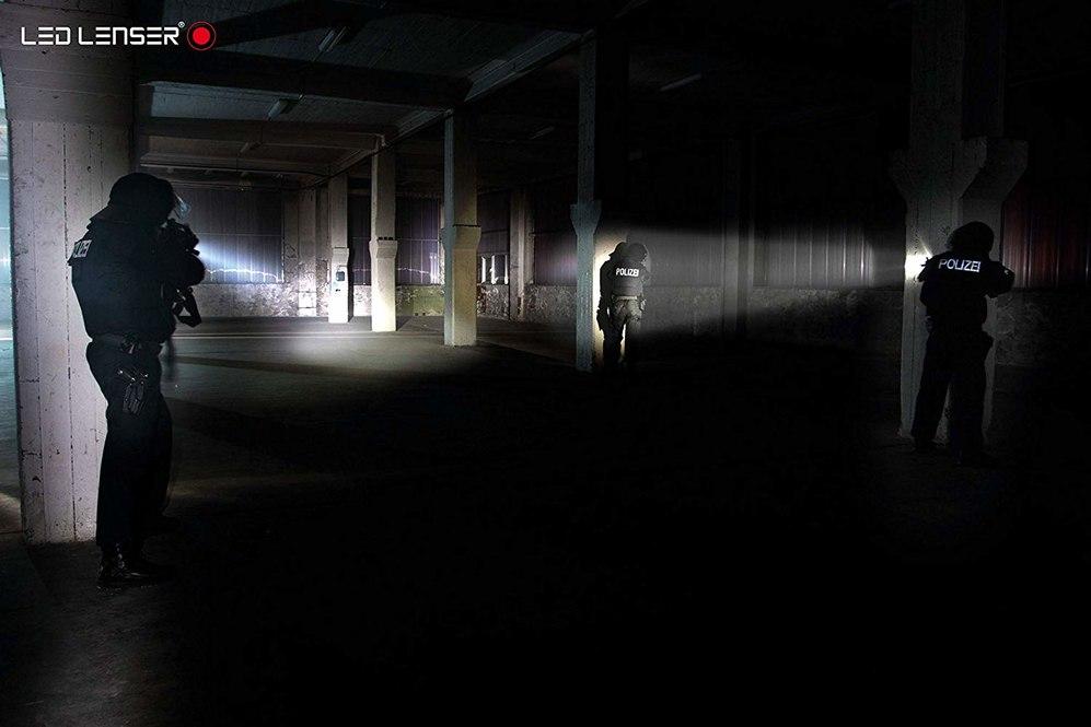 Drei Polizeibeamte stehen in einer dunklen, leeren Lagerhalle, leuchten mit ihren LEDLENSER Ledlenser Taschenlampe TT und halten Pistolen in der Hand. Der Strahl der langlebigen Ledlenser Taschenlampe TT erhellt teilweise den schattigen Raum. POLIZEI ist auf ihren Uniformen sichtbar.