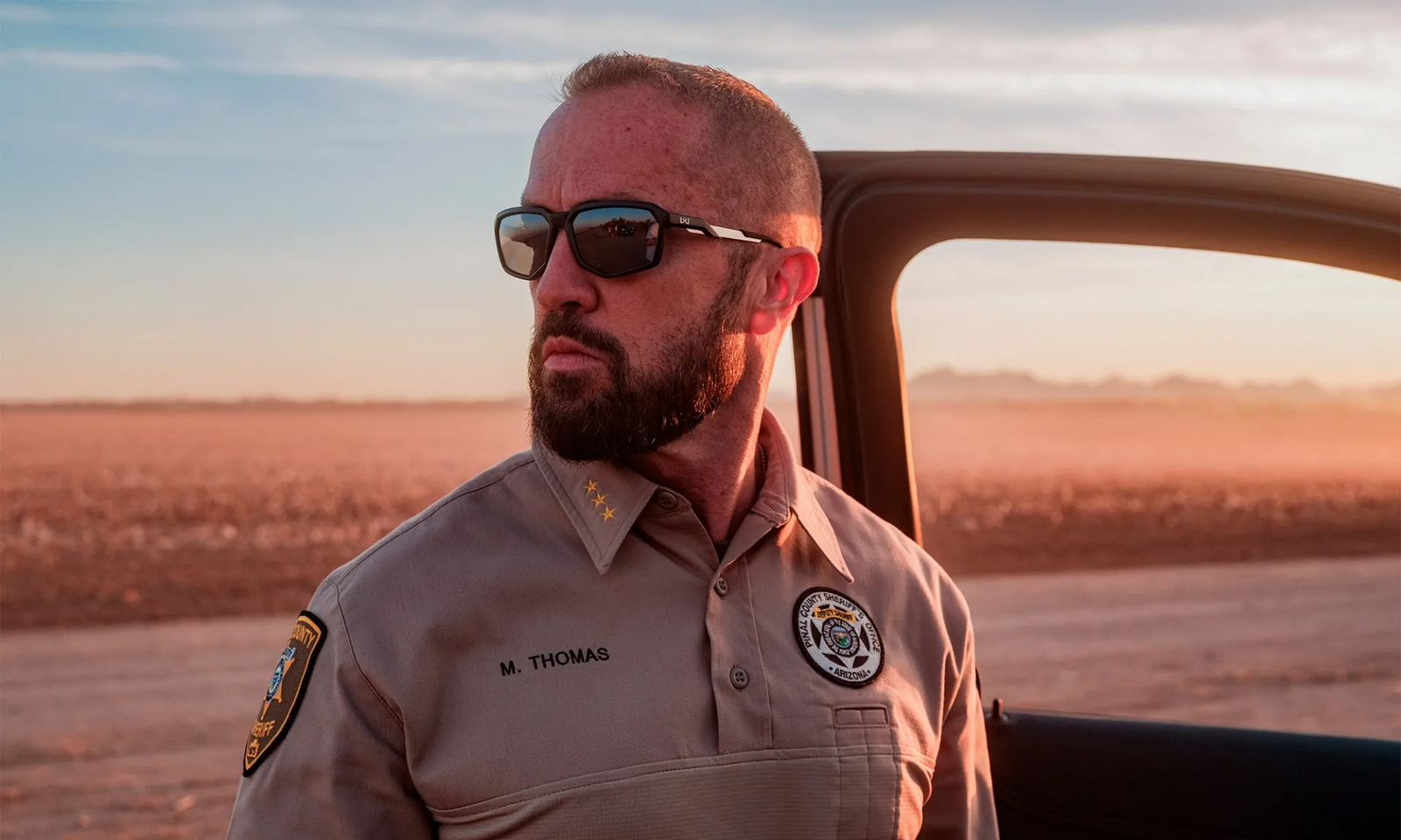 Ein bärtiger Mann in einer Sheriff-Uniform und einer WILEY X Wiley X Sonnenbrille RECON steht bei Sonnenuntergang neben einer offenen Autotür in der Wüste. Auf seinem Namensschild steht M. Thomas.