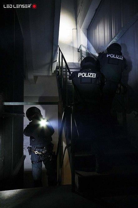 Drei bewaffnete Polizisten in taktischer Ausrüstung mit der Aufschrift Polizei führen eine Razzia in einem schummrigen Treppenhaus durch. Einer zielt mit der LEDLENSER Ledlenser Taschenlampe TT, während zwei mit ihren robusten Taschenlampen nach oben steigen.