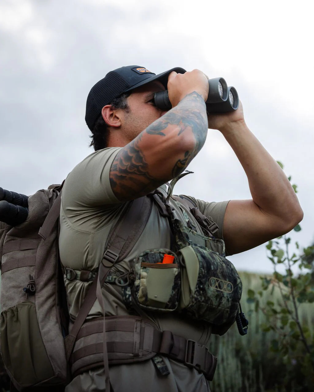 Eine tätowierte Person trägt einen Rucksack mit der EBERLESTOCK Recon Rangefinder Pouch. Sie trägt Outdoor-Ausrüstung und eine Mütze. Sie schaut durch ein Fernglas im Freien, im Hintergrund ist ein Gebüsch zu sehen.