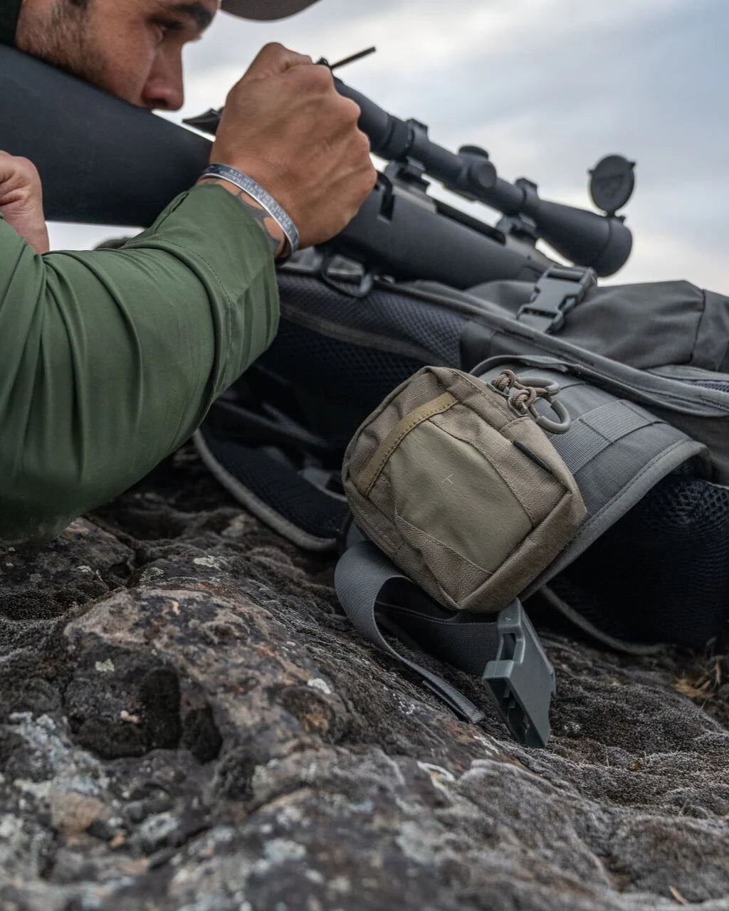 Eine Person in einem grünen langärmeligen Hemd zielt mit einem Gewehr auf felsigen Boden. In der Nähe liegen ein EBERLESTOCK Rucksack und eine Eberlestock Recon Utility Pouch, beide unter dem bewölkten Himmel.