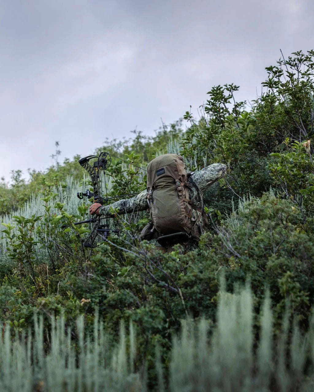 Eine in Tarnfarben gekleidete Person, die den ultraleichten EBERLESTOCK Vapor 2500 V2 Rucksack trägt, klettert unter einem bedeckten Himmel mit einem Compoundbogen auf einen buschigen, grünen Hügel und vermittelt ein raues Outdoor-Gefühl.