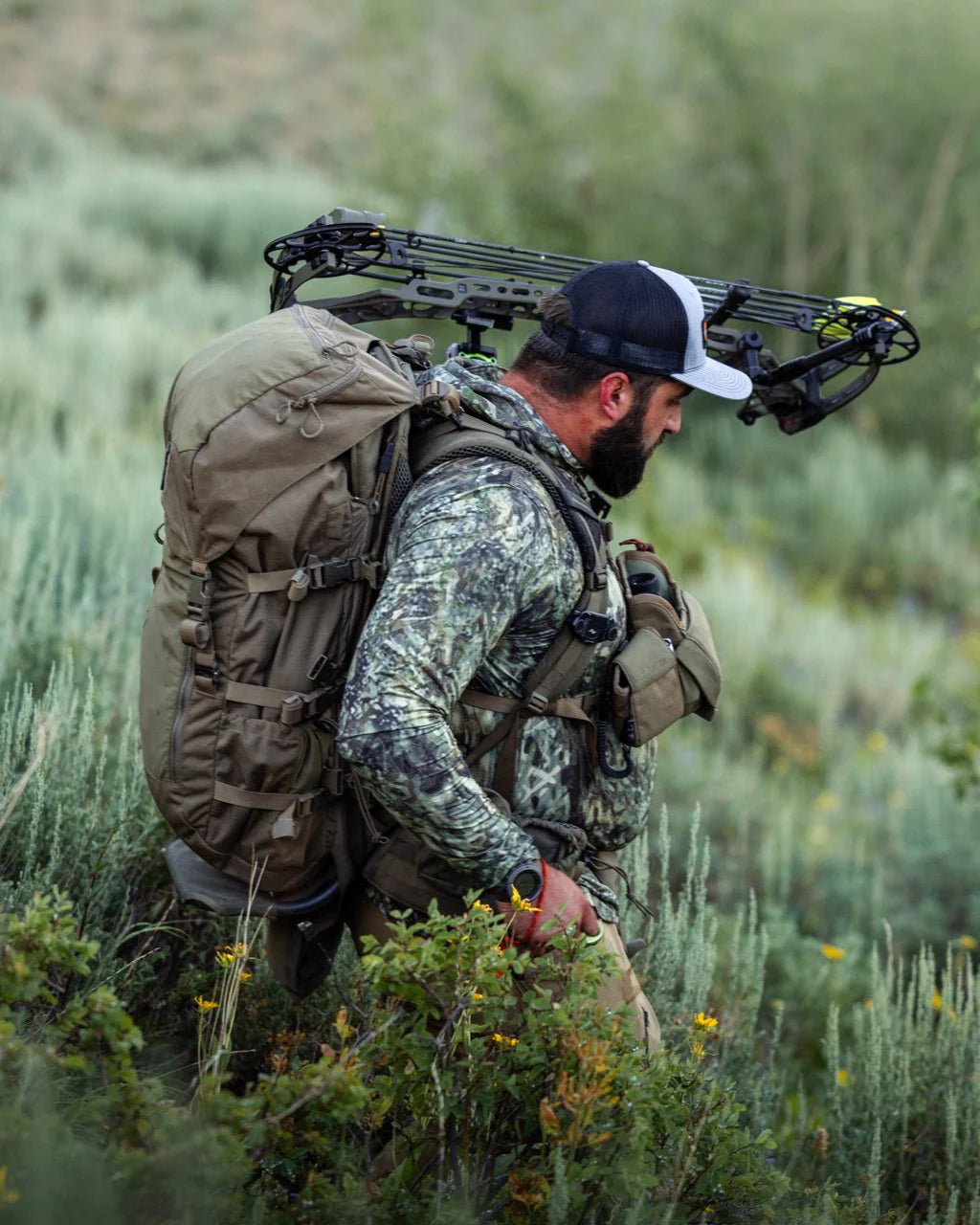 Ein Mann in Camouflage mit einem ultraleichten EBERLESTOCK Vapor 2500 V2 Rucksack geht durch grasbewachsenes Gelände und trägt einen Compoundbogen über der Schulter in der Wildnis.