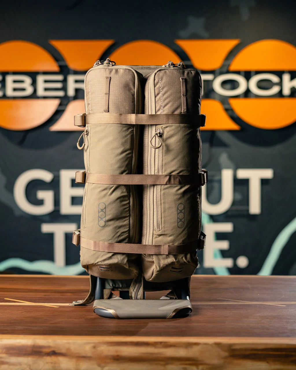 Der EBERLESTOCK Batwing V2, ein hellbrauner Rucksack mit mehreren Fächern und Gurten, wird auf einer Holzoberfläche gezeigt. Ein unscharfes "GET OUT THERE"-Schild und ein Teil des EBERLESTOCK-Namens erscheinen im Hintergrund und betonen seine EMOD-System-Kompatibilität.