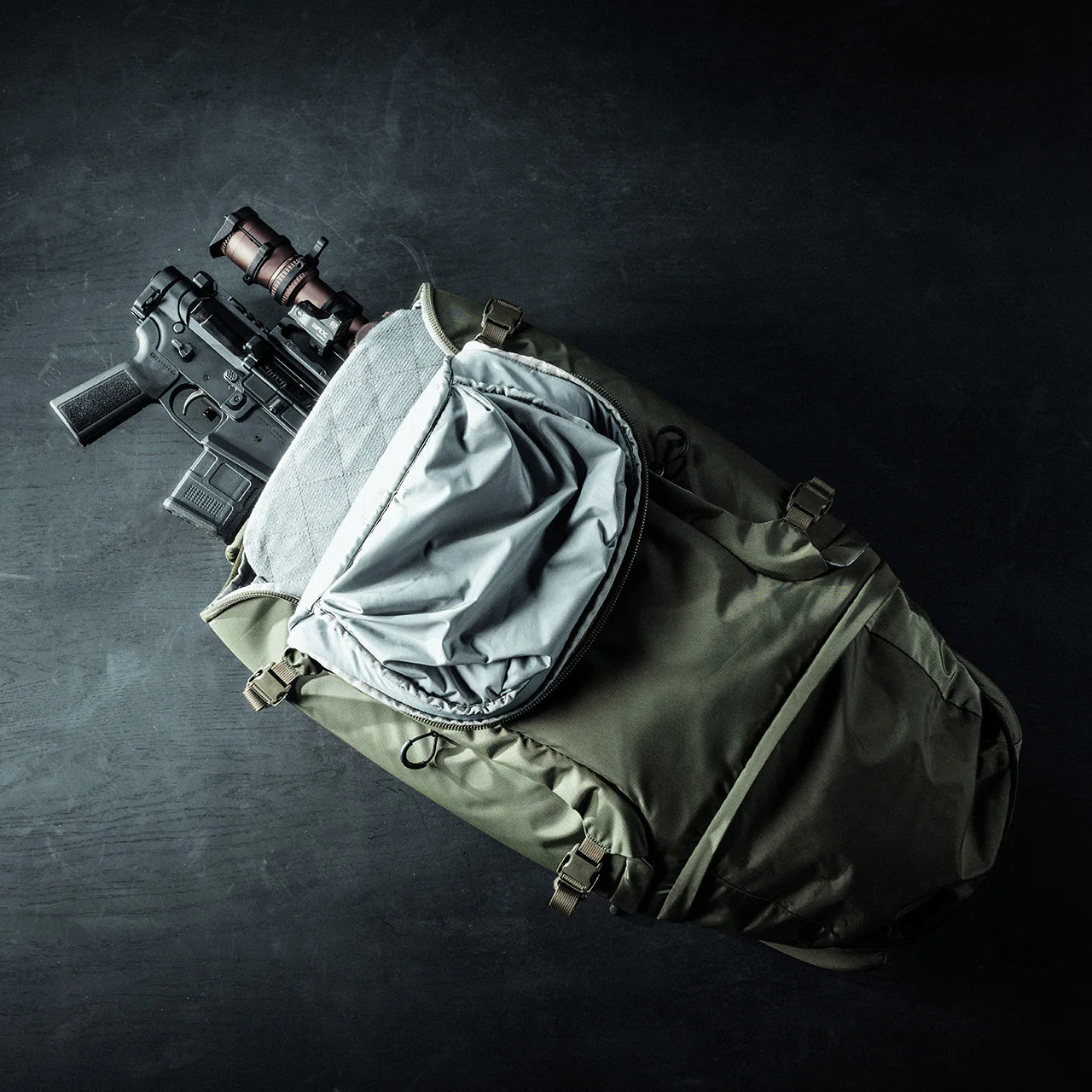 Ein EBERLESTOCK Eberlestock Fade Adapt liegt geöffnet auf einer dunklen Fläche und enthüllt ein schwarzes Gewehr mit einem braunen Zielfernrohr, das teilweise in das Hauptfach gepackt ist.