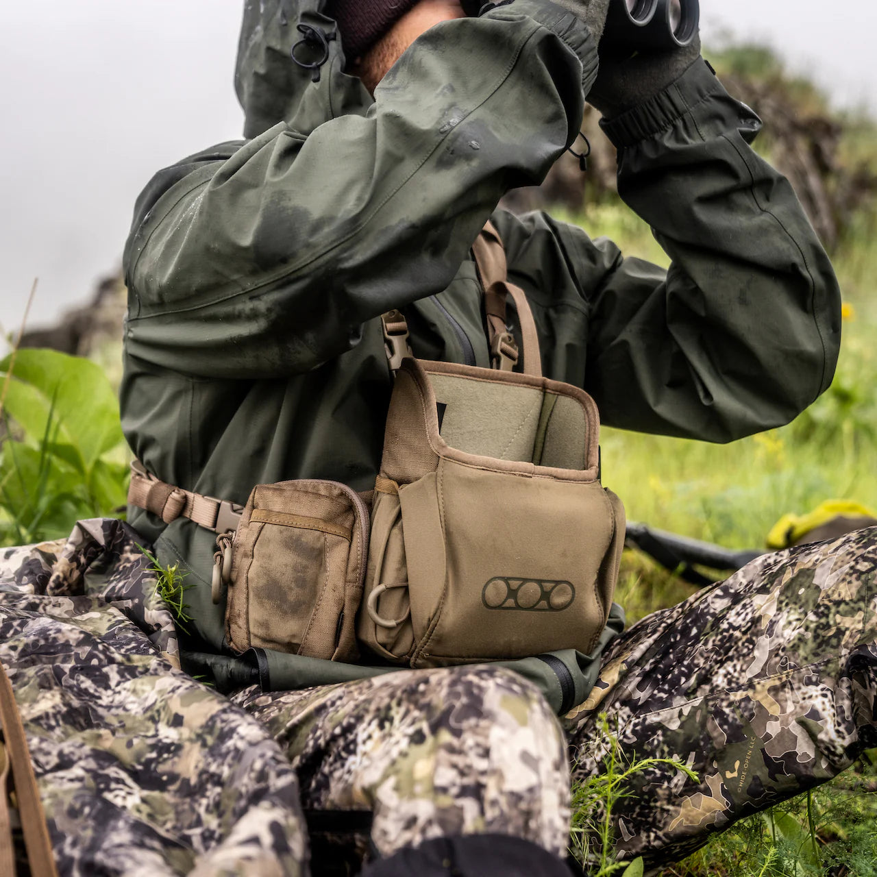 Eine Person in Camouflagekleidung sitzt auf Gras und hält ein Fernglas, sicher verstaut im EBERLESTOCK Recon Modular Bino Pack Small - die perfekte Wahl für echte Outdoor-Enthusiasten vor grüner Kulisse.