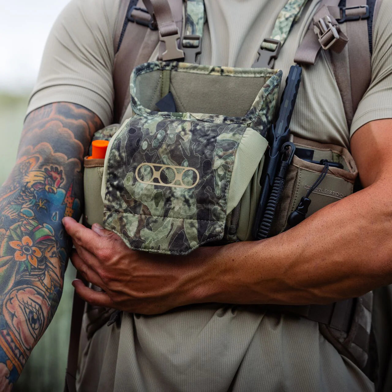 Eine Person mit einem farbenfrohen Unterarmtattoo trägt ein EBERLESTOCK Recon Modular Bino Pack Large, das mit Ausrüstung gefüllt ist - einem orangefarbenen Gegenstand, einem Multitool und einem Funkgerät - und stützt ihre Hand auf die Tasche dieses vielseitigen Fernglas-Tragesystems.