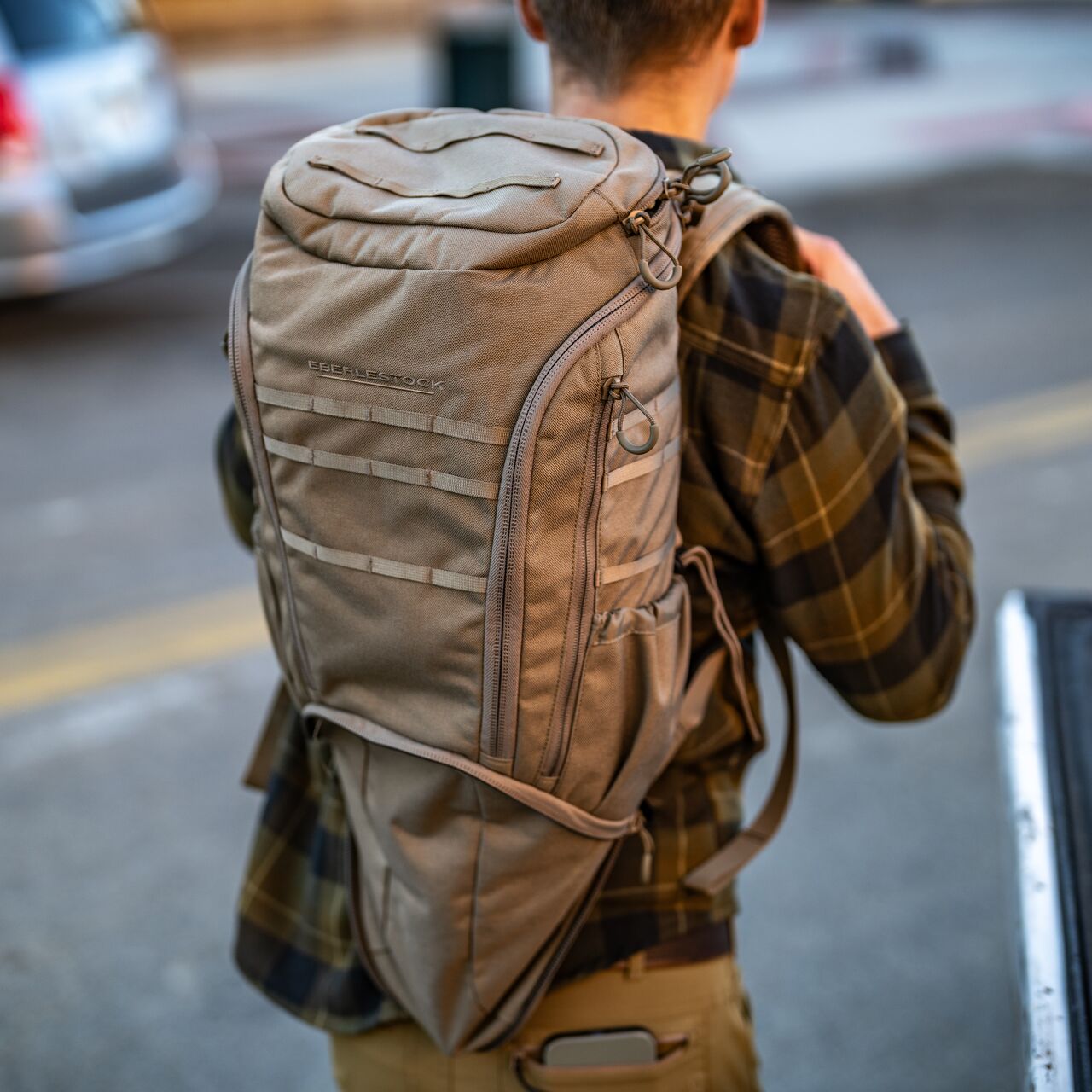 Eine Person in einem braunen karierten Hemd trägt das EBERLESTOCK Eberlestock Little Trick Pack mit mehreren Fächern und einer Tasche für verdeckte Waffen in einer städtischen Außenumgebung.