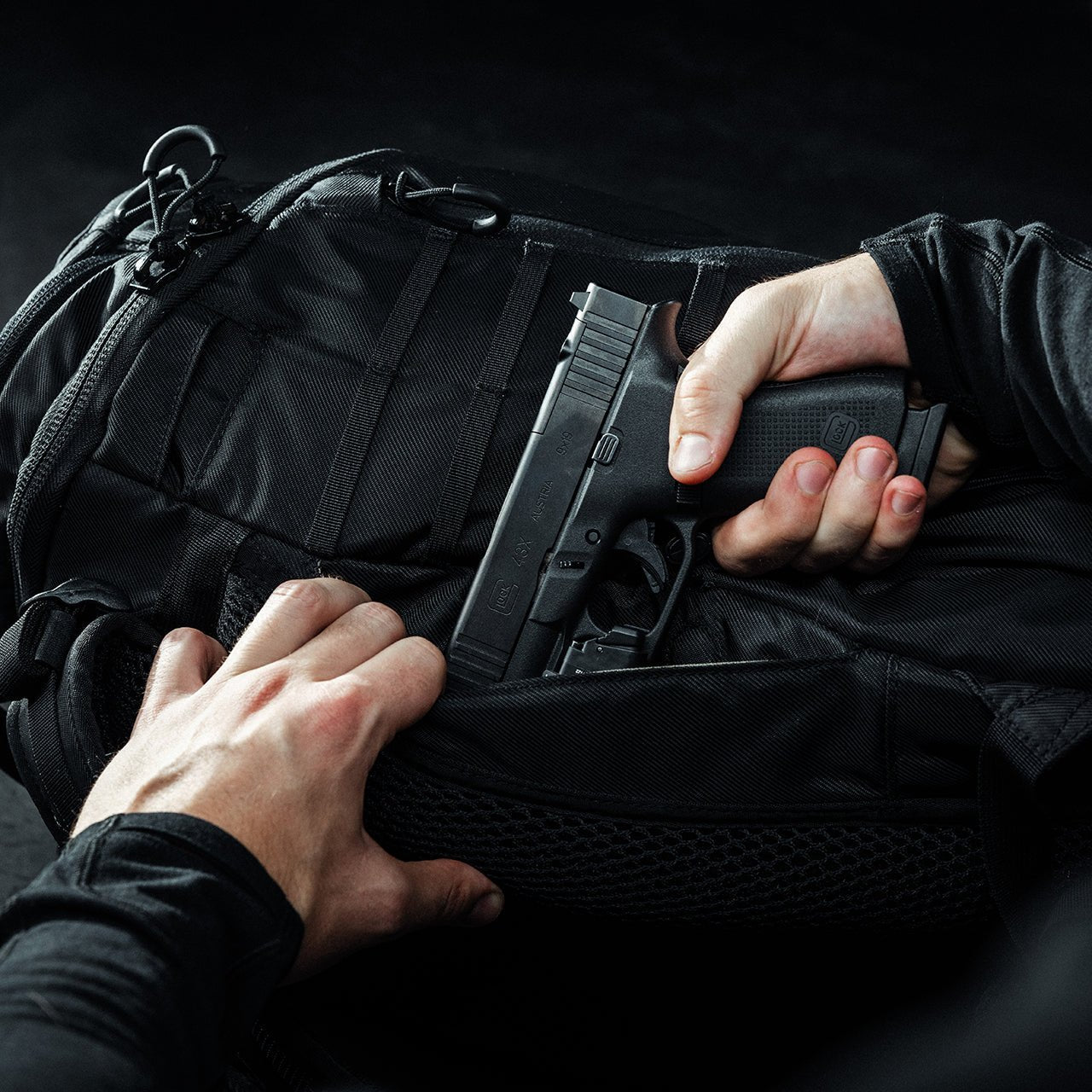 Eine Person in schwarzen Ärmeln legt eine schwarze Handfeuerwaffe in das versteckte Waffenfach eines schwarzen EBERLESTOCK Little Trick Pack vor einem dunklen Hintergrund.