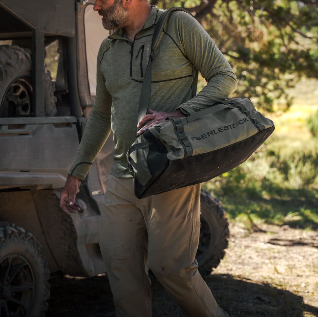 Ein Mann in einem militärgrünen langärmeligen Hemd und einer hellbraunen Hose trägt den EBERLESTOCK Hyllus Duffel 65 über der Schulter, während er im Freien neben einem Geländewagen steht und das Sonnenlicht durch die Bäume fällt.