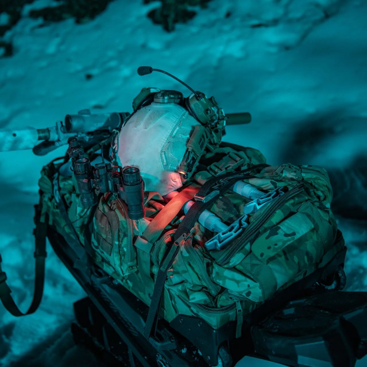 In einer verschneiten Szene, die von blauen und roten Lichtern beleuchtet wird, ruht ein Militärrucksack in Tarnfarben mit Ausrüstung, Helm, Taschenlampe und Funkgerät auf einem Schlitten - ergänzt durch den EBERLESTOCK Atlas Duffel.
