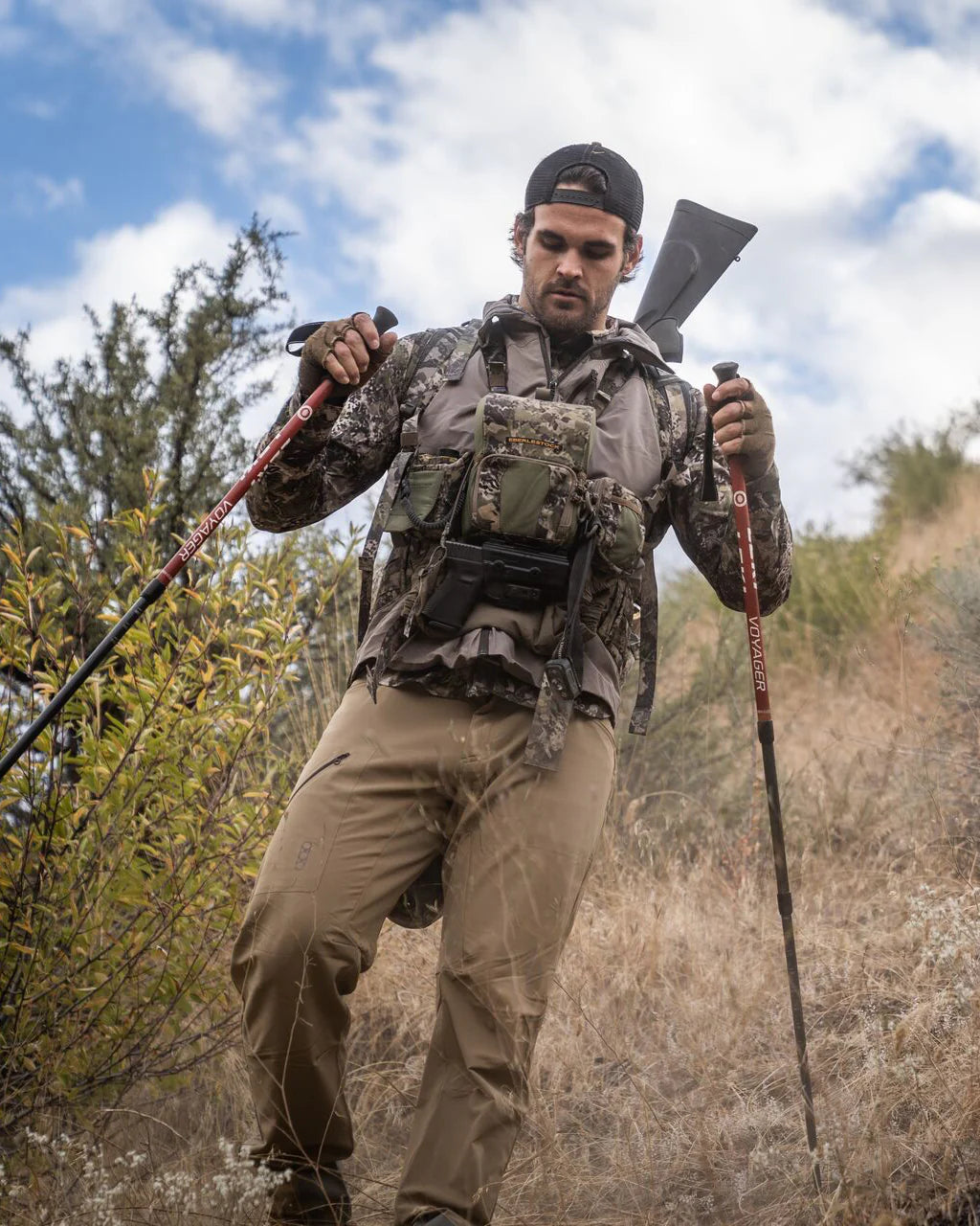 Ein Mann in Camouflage und mit umgekehrter Mütze wandert mit Trekkingstöcken, trägt ein Gewehr und ein EBERLESTOCK Recon MOLLE Panel auf der Brust. Der grasbewachsene Hang ist von Büschen gesäumt und der Himmel ist teilweise bewölkt.
