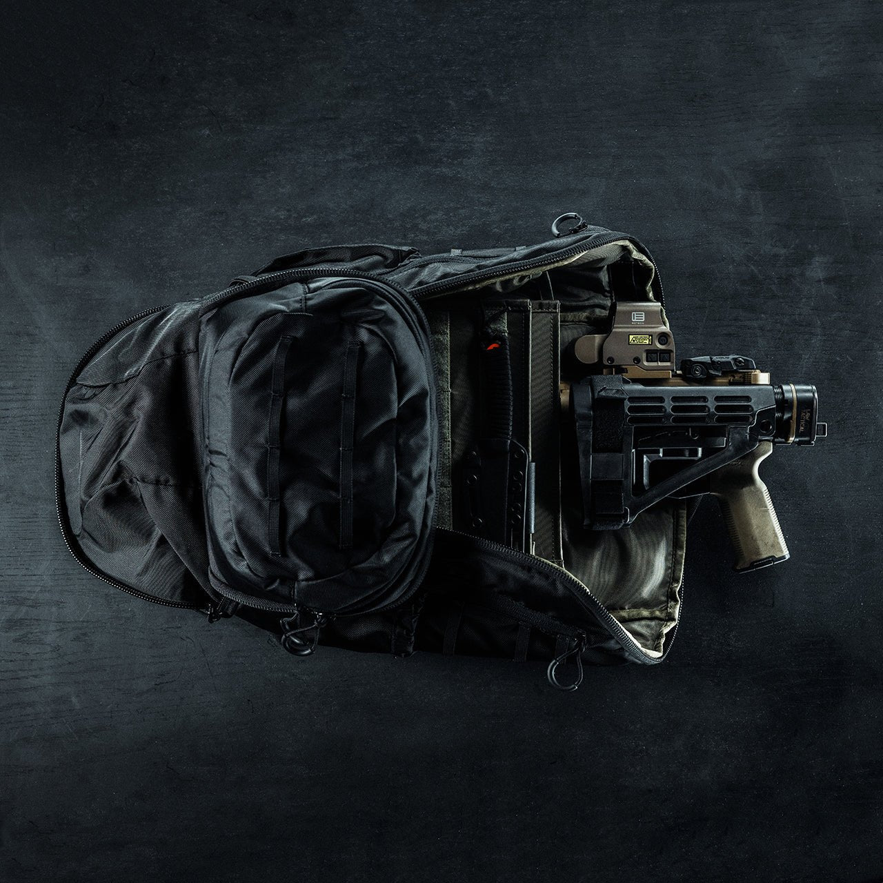 Ein schwarzer EBERLESTOCK Little Trick Pack liegt ohne Reißverschluss auf einer dunklen Fläche und offenbart eine kompakte Schusswaffe mit Zubehör und einem Magazin, das in seinem verdeckten Waffenfach verstaut ist.
