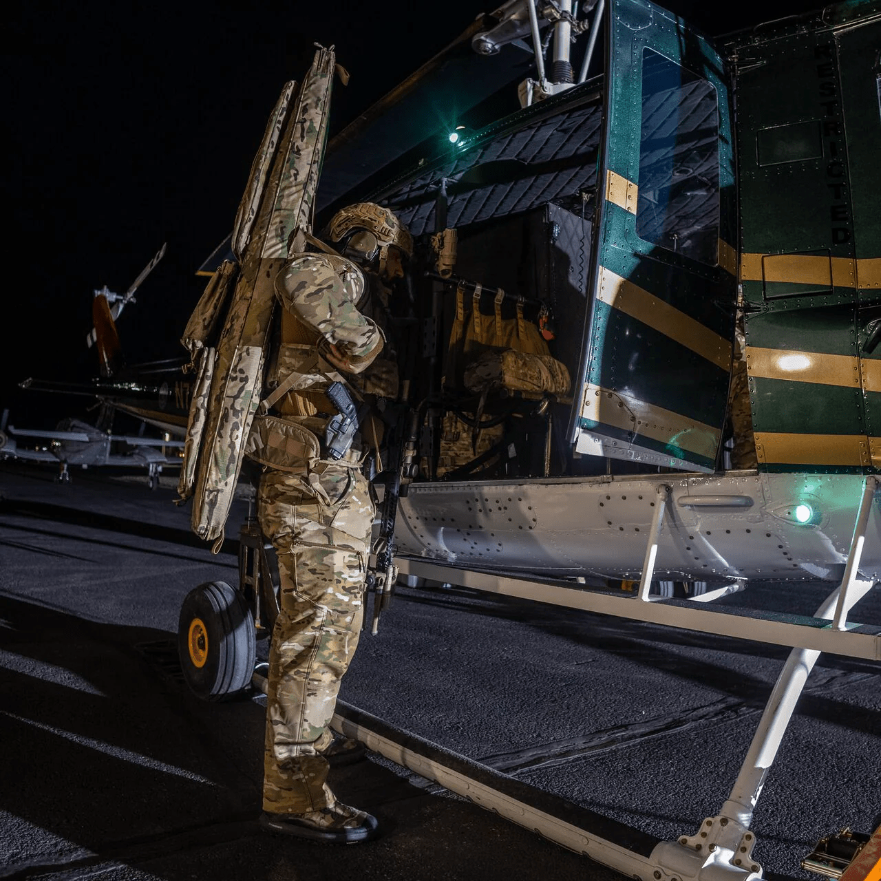 Ein Soldat in Tarnkleidung und mit Helm steht nachts neben einem Hubschrauber und hält eine EBERLESTOCK Sniper Sled Drag Bag 52" in der Hand, während die geöffnete Seitentür den Blick auf das schwach beleuchtete Rollfeld freigibt, und bereitet sich auf den Einstieg vor.