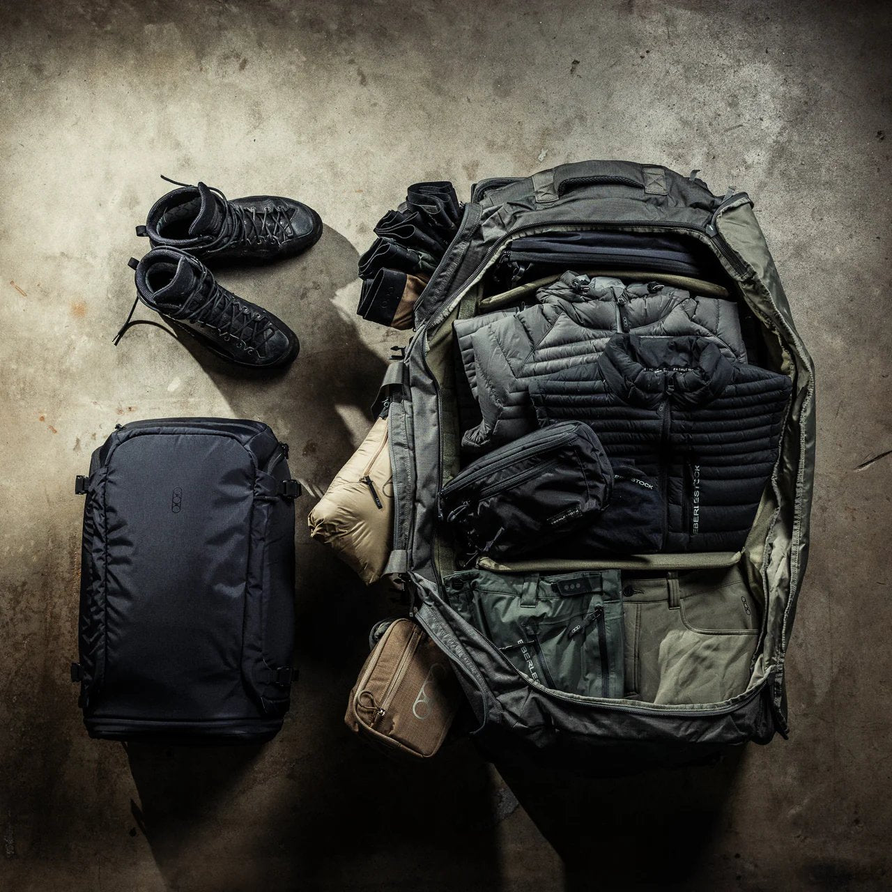 Ein EBERLESTOCK Atlas Duffel liegt offen auf dem Betonboden, fein säuberlich gepackt mit Kleidung, Jacken und Beuteln. Schwarze Stiefel und ein schwarzer Rucksack liegen daneben, alles organisiert für Reisen oder Outdoor-Abenteuer.