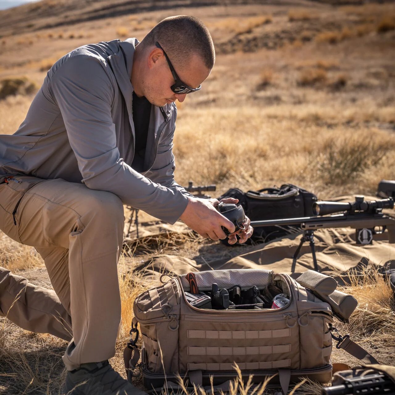 Ein Mann mit Sonnenbrille und Outdoor-Ausrüstung kniet im trockenen Gras und inspiziert die Kameraausrüstung neben einer geöffneten EBERLESTOCK Bang Bag. Neben ihm liegen zwei Gewehre und zusätzliche Ausrüstung, die an eine abgelegene Schießstand-Szene im rauen Sonnenlicht erinnern.