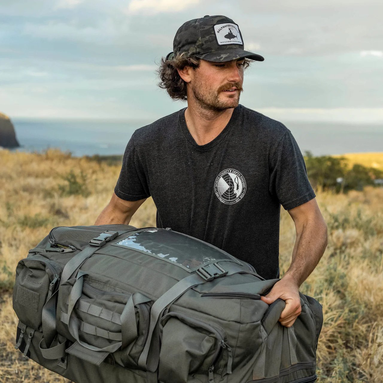 Ein Mann in dunklem T-Shirt und Tarnkappe steht auf einem trockenen Feld und hält die Eberlestock Atlas Duffel von EBERLESTOCK in der Hand. Im Hintergrund sind Hügel und ein teilweise bewölkter Himmel zu sehen - die ideale Umgebung, um diese Outdoor-Ausrüstung zu testen.