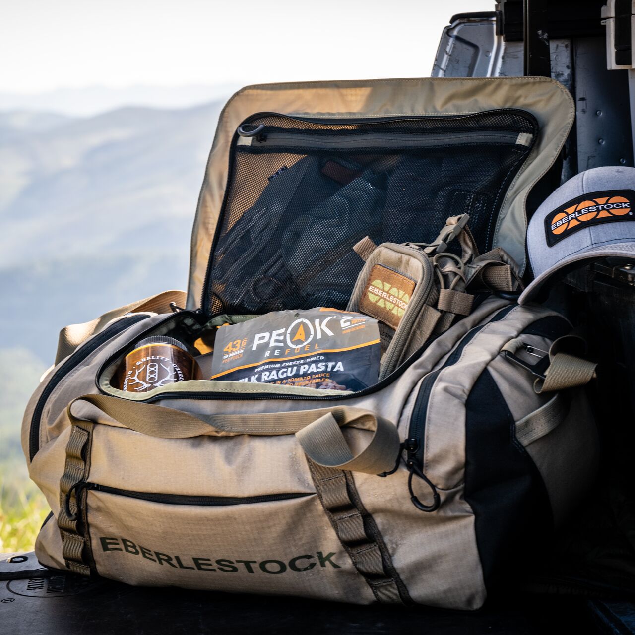 Ein EBERLESTOCK Hyllus Duffel 45 in Military Green liegt offen auf der Heckklappe eines Fahrzeugs, gefüllt mit Peak Refuel-Mahlzeiten und Outdoor-Ausrüstung, mit einer grauen EBERLESTOCK-Mütze in der Nähe. Unscharfe Berge bilden den malerischen Hintergrund.