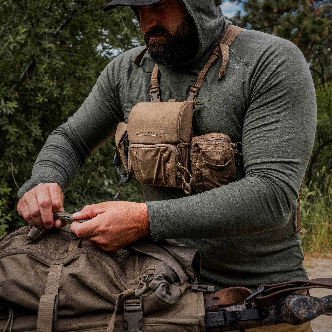 Eine Person, die ein grünes Kapuzenshirt trägt, justiert im Freien die Ausrüstung an einem hellbraunen Rucksack. Sie trägt ein EBERLESTOCK Recon Modular Bino Pack Small mit mehreren Taschen und hat ein Gewehr mit Tarnmuster umgehängt. Im Hintergrund sind Bäume zu sehen.