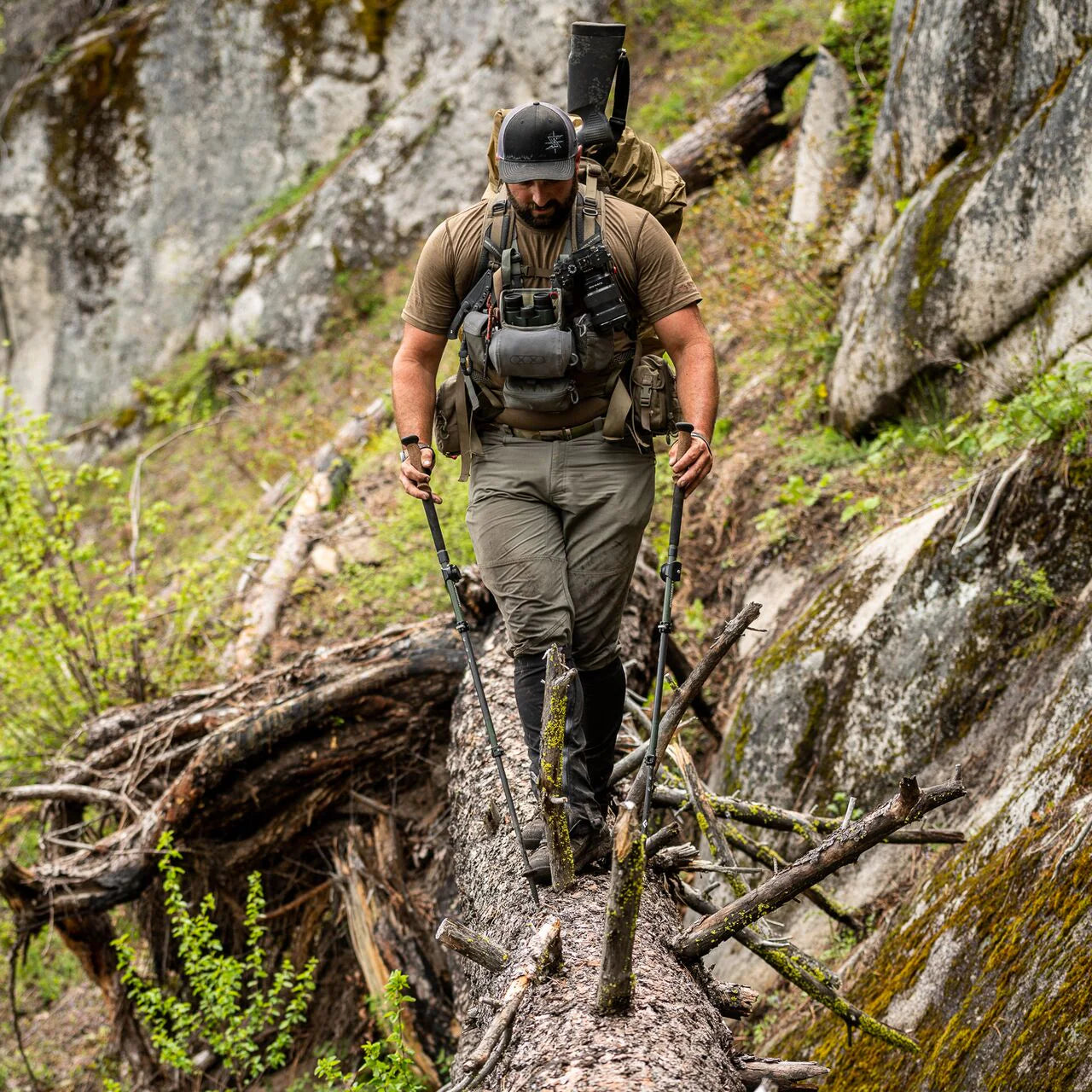 Ein Mann in Outdoor-Ausrüstung, perfekt für Outdoor-Liebhaber, überquert einen umgestürzten Baumstamm mit Trekkingstöcken, ein EBERLESTOCK Recon Modular Bino Pack Small auf der Brust, vor einem felsigen Waldhang.