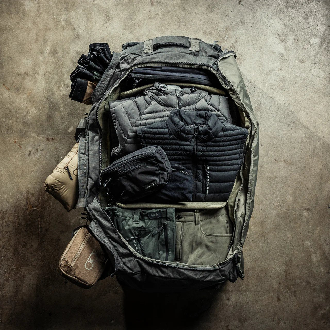Ein EBERLESTOCK Atlas Duffel liegt offen auf Beton, ordentlich gepackt mit Jacken, Hosen und Reisetaschen - ein Beispiel dafür, wie dieser vielseitige Seesack Kleidung und Ausrüstung ordnet.