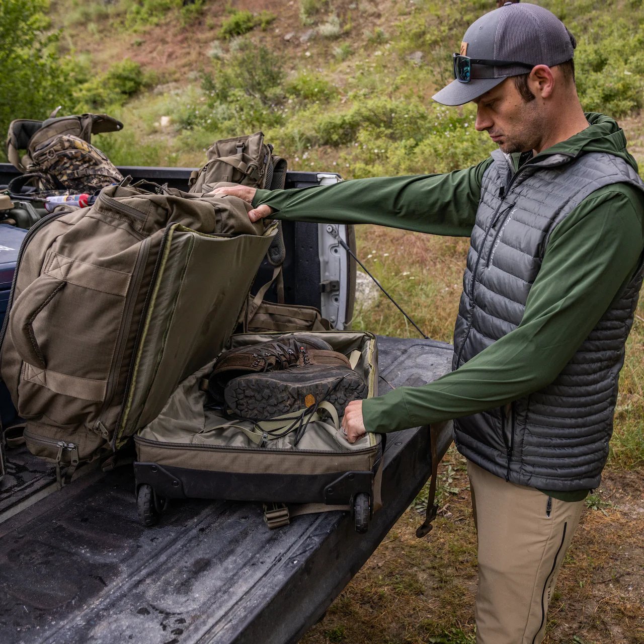 Ein Mann in Outdoor-Ausrüstung packt Wanderschuhe in einen EBERLESTOCK Atlas Duffel auf der Heckklappe eines Pickups, umgeben von Rucksäcken, Ausrüstung und einem Rollkoffer in einer grasbewachsenen, bewaldeten Umgebung.