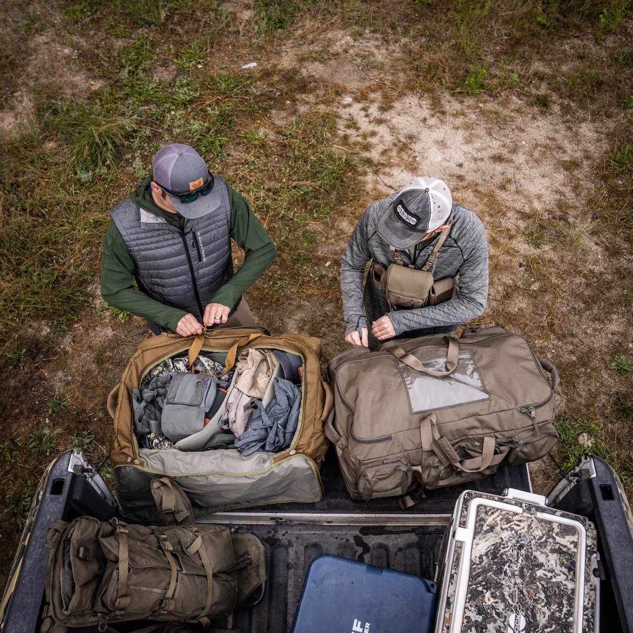Zwei Personen in Outdoor-Kleidung stehen an der offenen Heckklappe eines Lastwagens und packen einen mit Ausrüstung gefüllten EBERLESTOCK Atlas Duffel auf grasbewachsenem Boden ein oder aus, von oben gesehen.