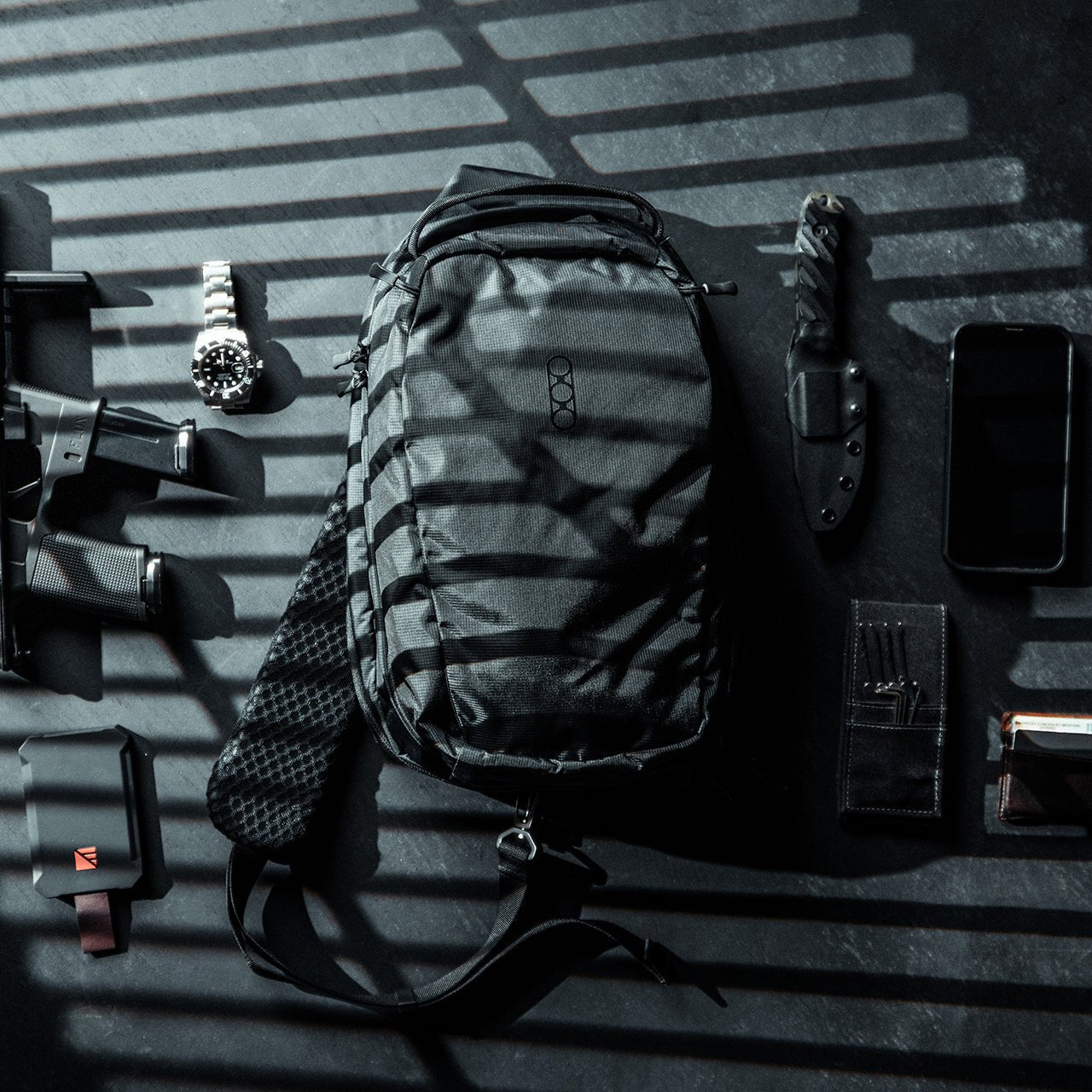 Eine EBERLESTOCK Eberlestock Fade Sling taktische Tasche in Schwarz liegt auf einer dunklen Fläche, ordentlich arrangiert mit einer Uhr, einem Messer, einer Handfeuerwaffe, einem Magazin, einem Smartphone, einer Brieftasche und einem kleinen Gerät unter dramatischer, gestreifter Beleuchtung.