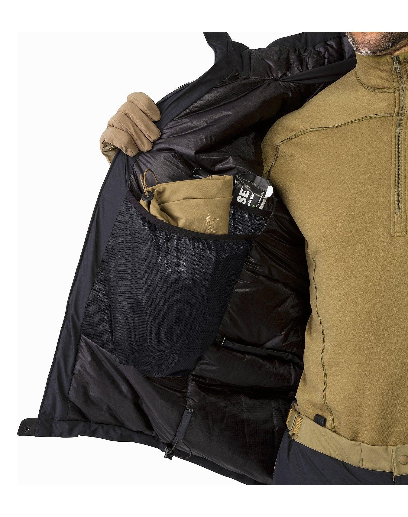 Eine Person in hellbrauner Outdoor-Ausrüstung öffnet den Reißverschluss einer schwarzen ARC'TERYX LEAF Arc'teryx Cold WX Jacket SV Gen1.4 und zeigt eine innere Netztasche mit hellbraunen Handschuhen und kleinen Gegenständen; nur der Torso und der rechte Arm sind sichtbar.