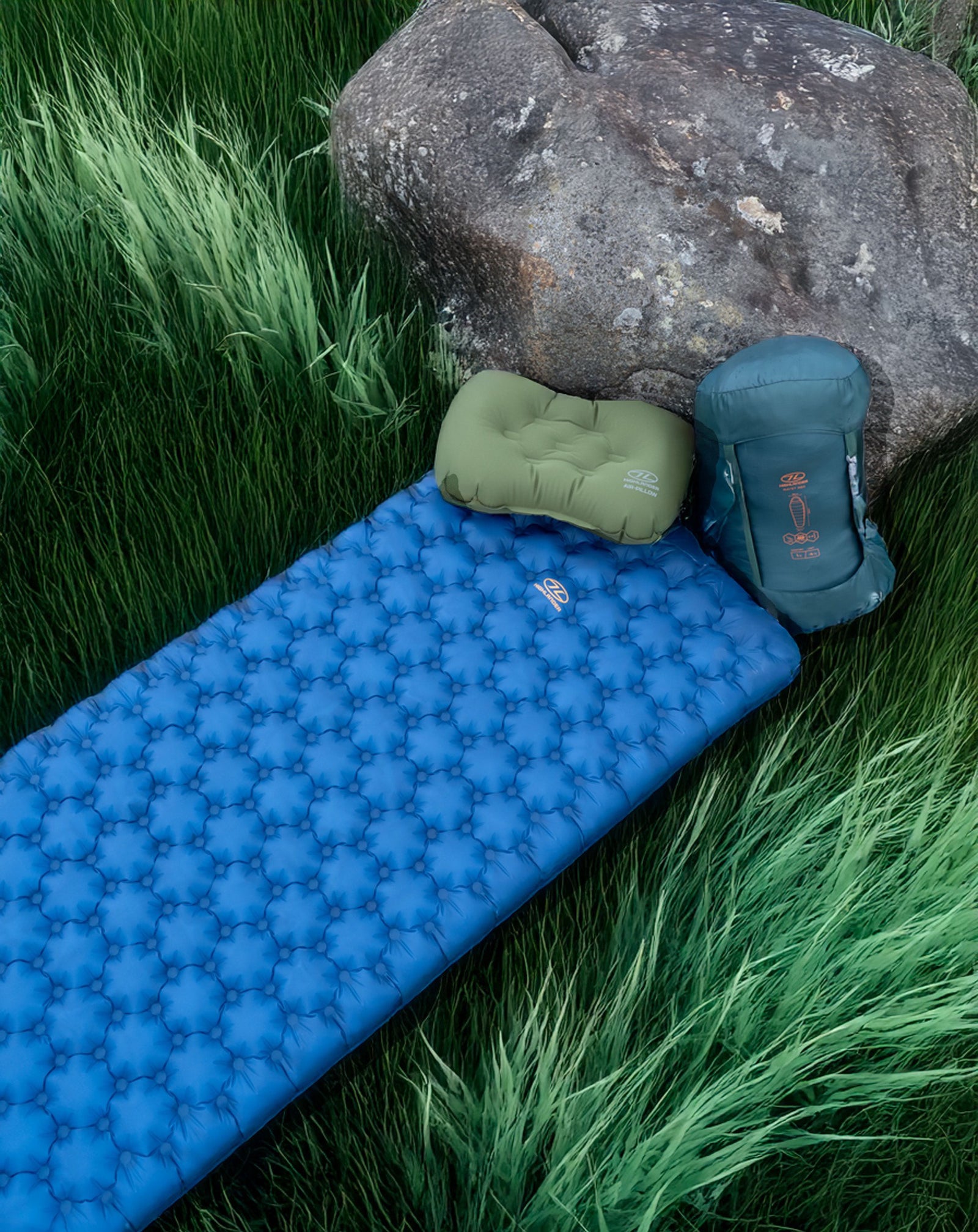 Eine Highlander NAP PAK ARCTIC AIR MAT von HIGHLANDER FORCES, ein grünes aufblasbares Kissen und ein verpackter grüner Schlafsack liegen im hohen Gras neben einem großen Felsen.