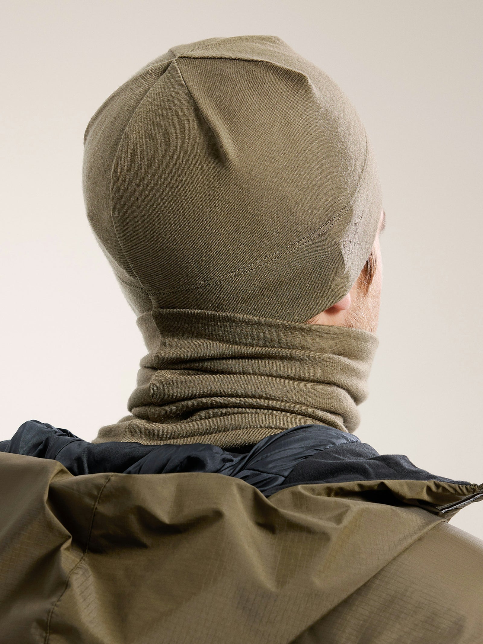 Eine Person in einer khakifarbenen Arc'teryx Cold WX Neck Gaiter AR von ARC'TERYX LEAF und passender Mütze, von hinten gesehen mit einer olivgrünen Jacke vor einem neutralen Hintergrund, ideal für kaltes Wetter.