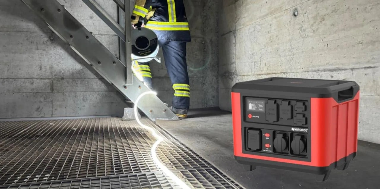 Ein Feuerwehrmann in Uniform steigt mit einem Schlauch eine Metalltreppe hinauf, während im Vordergrund eine NORDRIDE POWER-X Power Station 2000W für unterbrechungsfreie Stromversorgung am Boden sorgt.