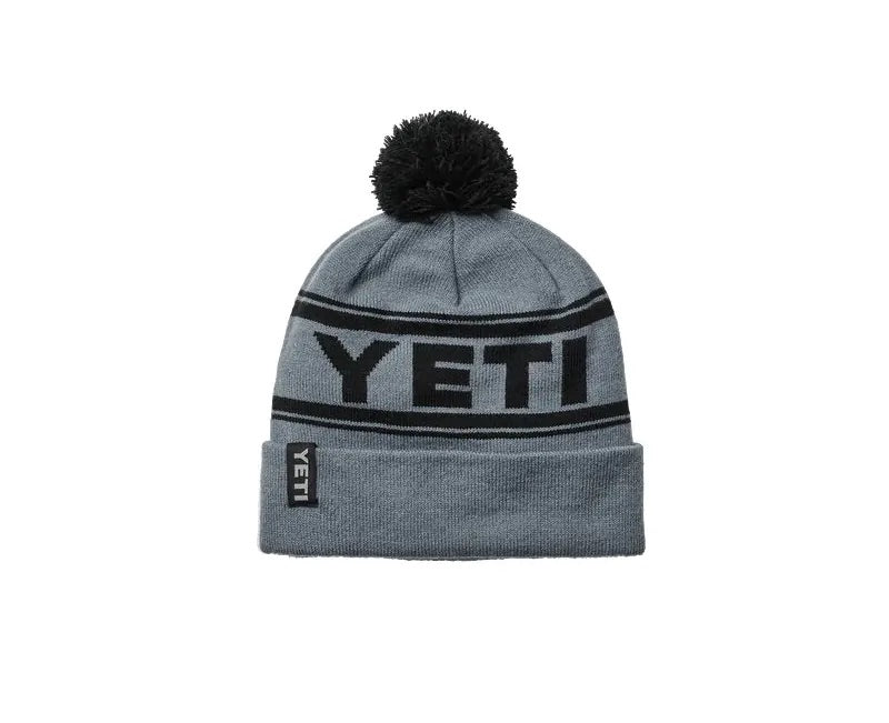 Die YETI® Logo Retro Knit Beanie von YETI ist eine graue Mütze mit einem schwarzen Bommel, einem auffälligen schwarzen YETI-Schriftzug und einem kleinen YETI-Anhänger an der Krempe - die perfekte warme Mütze für kalte Tage.