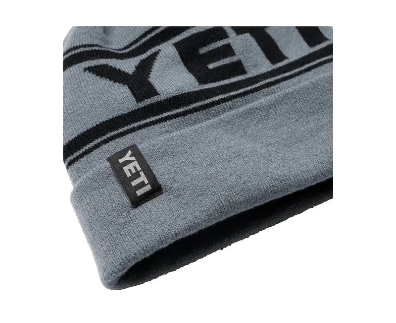 Eine Nahaufnahme der grauen YETI® Logo Retro Knit Beanie, einer warmen Mütze mit auffälligem YETI-Schriftzug und einem kleinen schwarzen Stoffanhänger mit der Aufschrift YETI an der gefalteten Krempe.