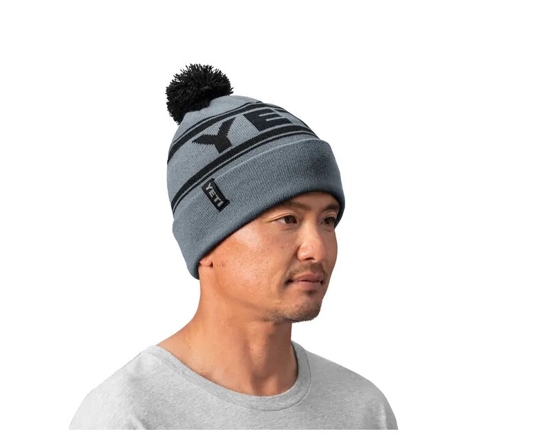 Ein Mann mit einer hellblauen YETI® Logo Retro Knit Beanie mit schwarzer Bommel und fettem Logoschriftzug, gepaart mit einem hellgrauen Rundhalsshirt, schaut leicht nach links.