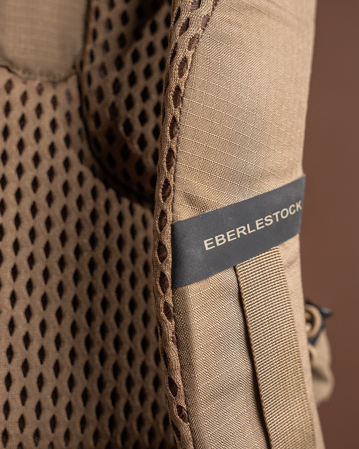 Nahaufnahme eines gepolsterten, hellbraunen Rucksackriemens mit Mesh-Details und dem EBERLESTOCK-Logo auf einem Stoffanhänger, der das robuste Design des Eberlestock Brute Scout von EBERLESTOCK unterstreicht.