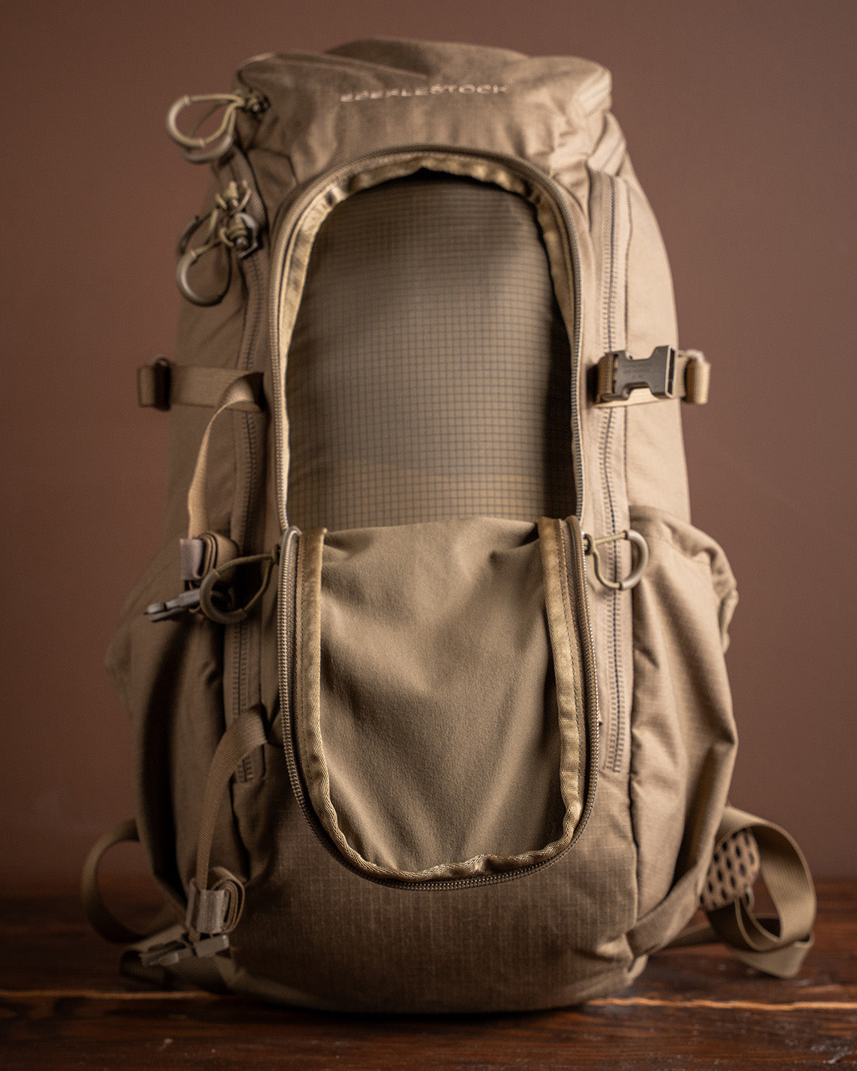 Der taktische Rucksack EBERLESTOCK Brute Scout in tan/coyote brown aus Ripstop-Gewebe mit Reißverschlussfächern, Schnallenriemen und D-Ringen ist aufrecht auf einer Holzfläche mit braunem Hintergrund abgebildet.