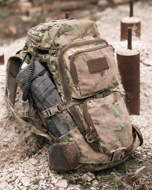 Ein EBERLESTOCK Eberlestock FAC Track - Military Green taktischer Rucksack mit großen Reißverschlusstaschen und einer Netztasche an der Seite sitzt auf felsigem Boden und hält ein aufgerolltes schwarzes Seil und einen zylindrischen Gegenstand; dahinter befinden sich braune Metallstangen.