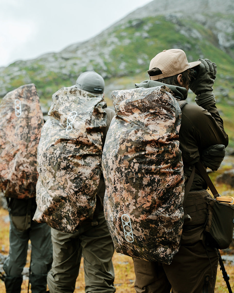 Drei Wanderer mit großen EBERLESTOCK Rucksäcken, geschützt durch die wendbare Eberlestock Rucksack-Regenhülle Large - Mountain / Blaze Orange, stehen im Gebirge. Einer sieht durch Fernglas. Alle tragen Outdoor-Kleidung und Caps, von hinten fotografiert.