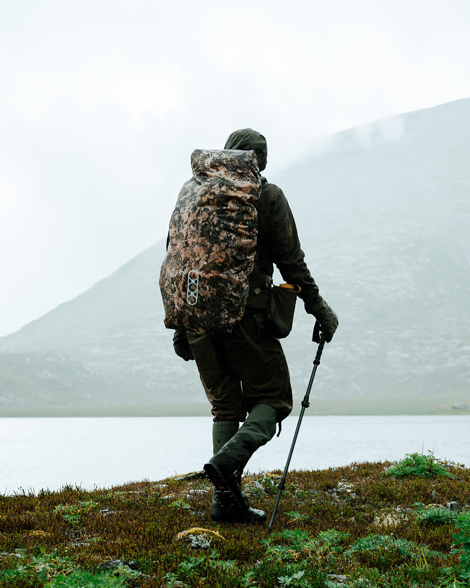 Eine Person in Outdoor-Ausrüstung trägt einen EBERLESTOCK Rucksack mit einer großen wendbaren Rucksack-Regenhülle (Mountain/Blaze Orange), steht auf einem grasbewachsenen Seegrund mit nebligen Bergen im Hintergrund und hält einen Wanderstock.