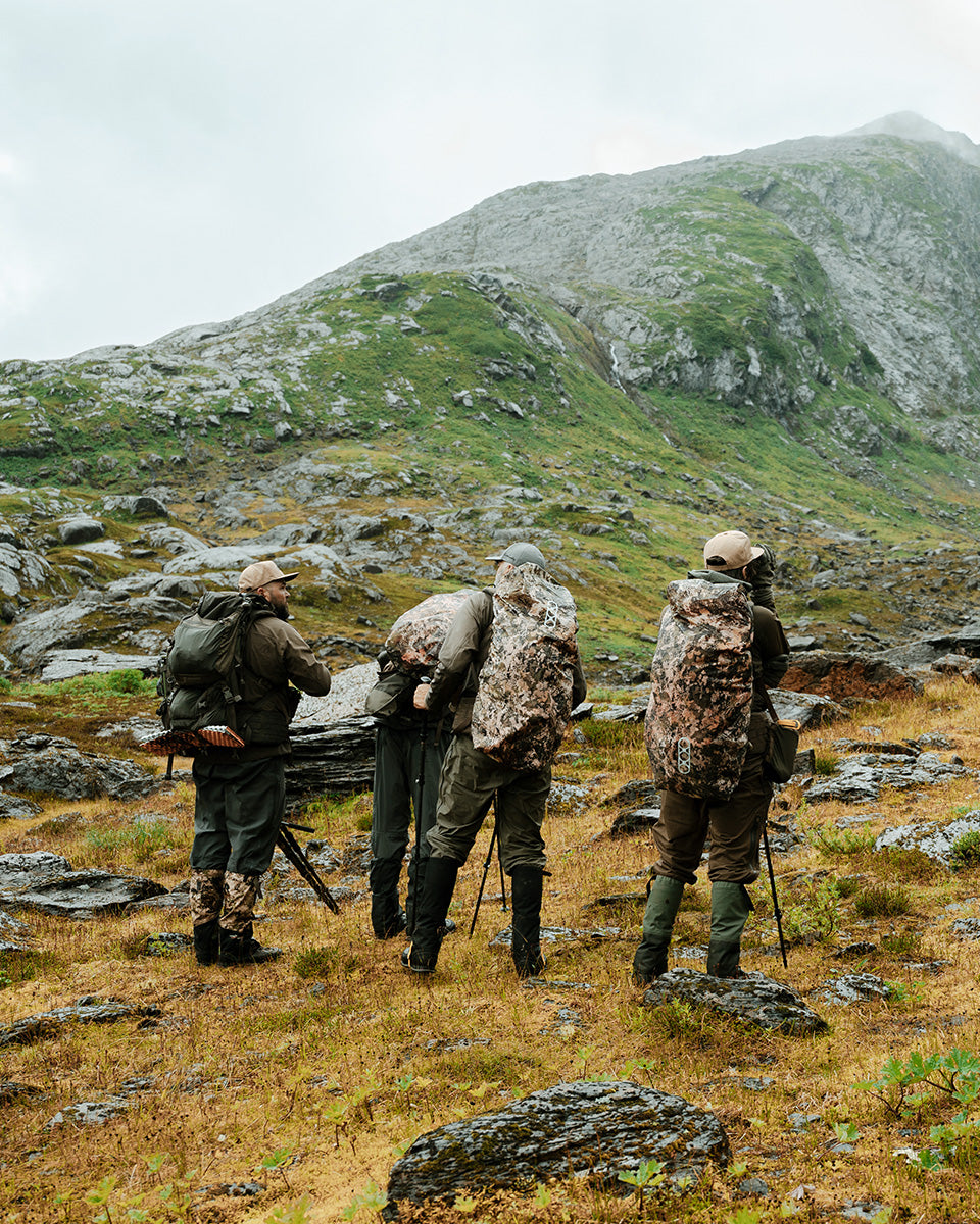 Vier Personen in Camouflage stehen mit Stativen und Rucksäcken - ausgestattet mit der EBERLESTOCK Rucksack-Regenhülle Large wendbar - Mountain / Blaze Orange - auf einem felsigen Hügel und blicken auf einen grünen Berg unter wolkenverhangenem Himmel.