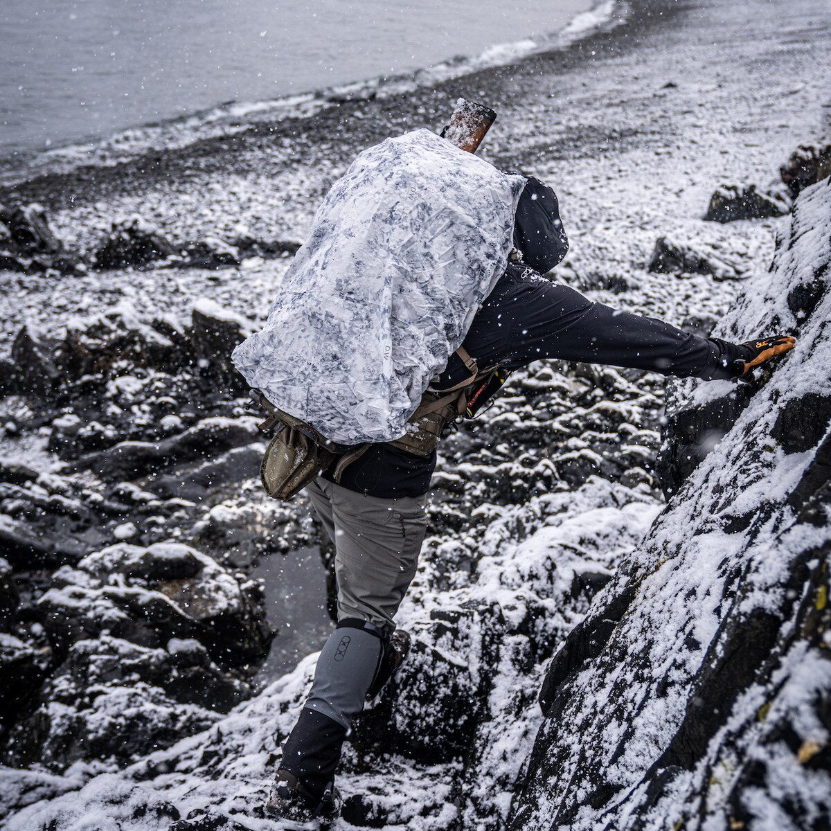 In Winterkleidung und mit einer EBERLESTOCK Rucksack-Regenhülle Large wendbar - Coyote / D. Winter steigt eine Person einen verschneiten, felsigen Hang am Ufer hinauf, während um sie herum Schnee fällt.
