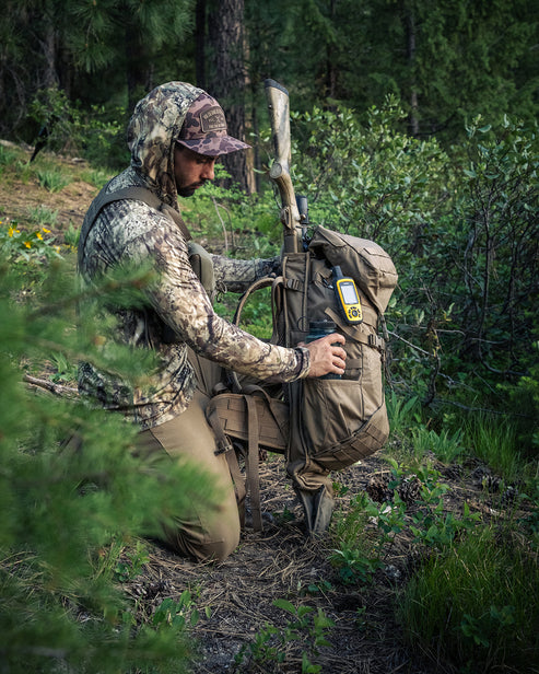 Eine Person in Tarnkleidung kniet in einem Wald und richtet einen EBERLESTOCK Gunrunner Pack - Dry Earth mit einem Gewehr und einem gelben GPS-Gerät in einer Außentasche aus.