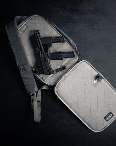 Eine schwarze Schusswaffe mit erweitertem Magazin ruht in einer offenen grauen EBERLESTOCK Eberlestock Fade Sling XL Tasche mit Riemen und Innenetikett, die sich perfekt für den Transport von EDC-Ausrüstung vor einem dunklen Hintergrund eignet.