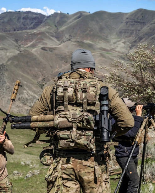 Eine Person in Tarnfarben und mit einer grauen Mütze trägt in den Bergen eine taktische Ausrüstung, darunter eine EBERLESTOCK Rip-Away Medical Pouch small am Gürtel und ein Gewehr auf dem Rücken. Andere stehen in der Nähe, einer mit einer Kamera.