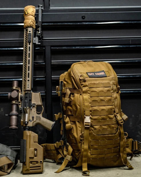 Ein hellbraunes EBERLESTOCK Mission Assault Pack mit modularem Design, mehreren Gurten und einem "NOT YOURS"-Aufnäher steht neben einem passenden hellbraunen Gewehr mit Zielfernrohr auf einer schwarzen Metalloberfläche.