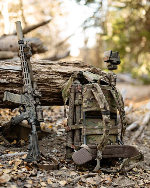 Ein getarntes Gewehr ruht auf dem EBERLESTOCK Mission EMOD Frame in einem Wald, umgeben von herabgefallenen Blättern und Baumstämmen im gedämpften Tageslicht - und unterstreicht die hochwertige taktische Ausrüstung.