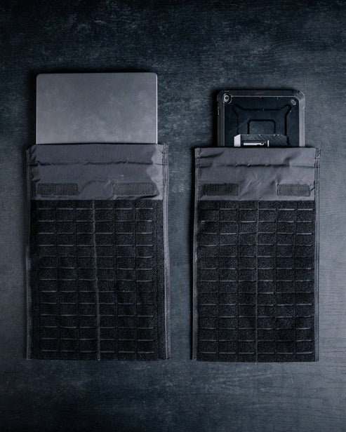 Zwei schwarze taktische Taschen, darunter die EBERLESTOCK Fade Laptop Blocker Sleeve, ruhen auf einer dunklen Oberfläche - eine hält einen Laptop, die andere ein elektronisches Gerät; beide sind teilweise sichtbar und bieten optimalen Laptopschutz.