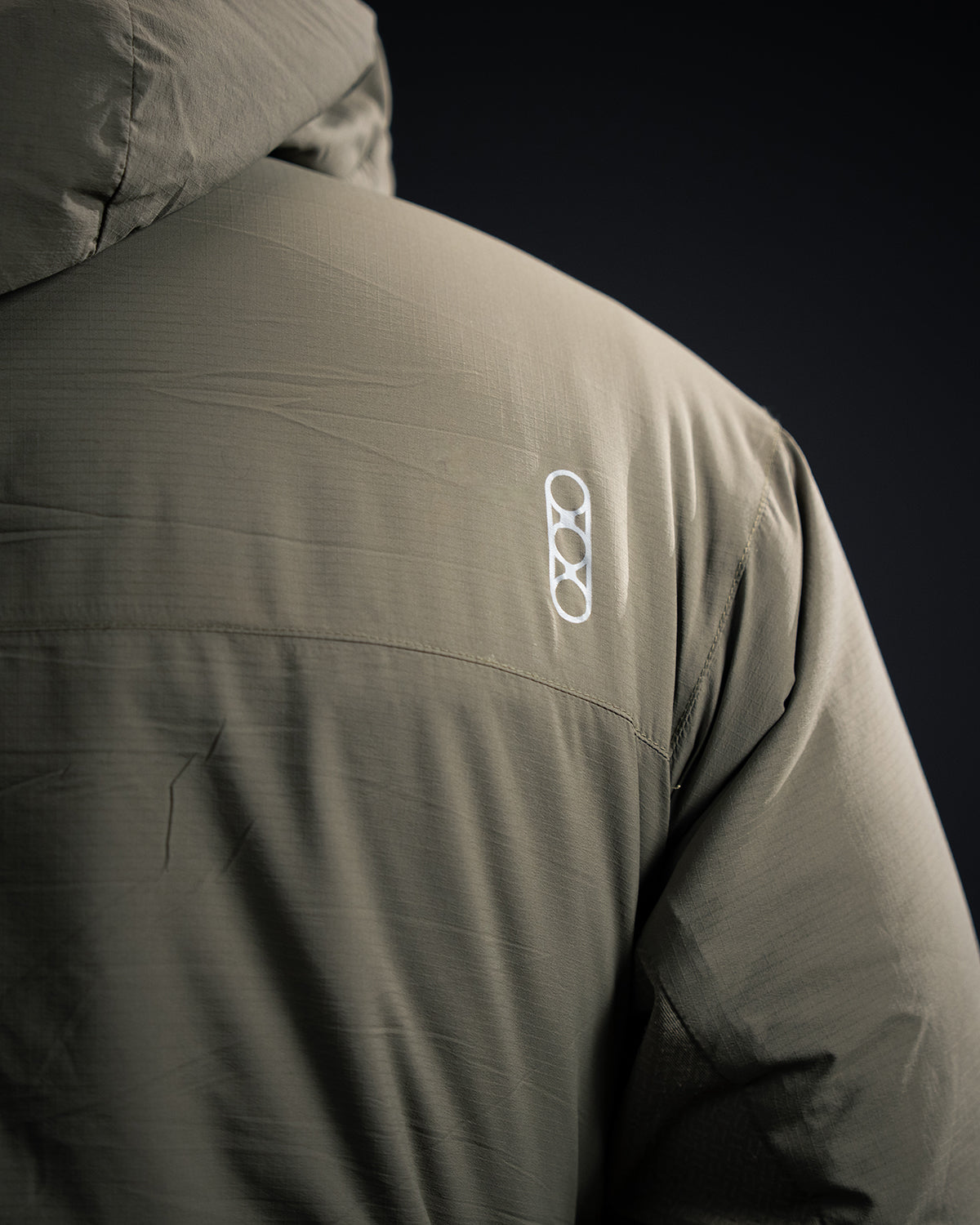 Nahaufnahme des oberen Rückens der EBERLESTOCK Alturas Hooded Jacket - Ranger Green, die ein kleines weißes geometrisches Logo mit überlappenden Kreisen zeigt. Der Stoff hat ein subtiles, strukturiertes Aussehen.