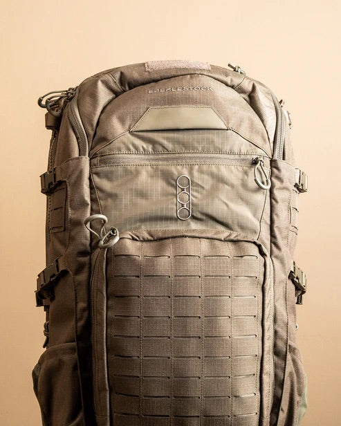 Der EBERLESTOCK Spectre, ein großer olivgrüner Outdoor-Rucksack mit mehreren Reißverschlüssen, Schnallen und Außenriemen, steht aufrecht vor einem schlichten beigen Hintergrund.